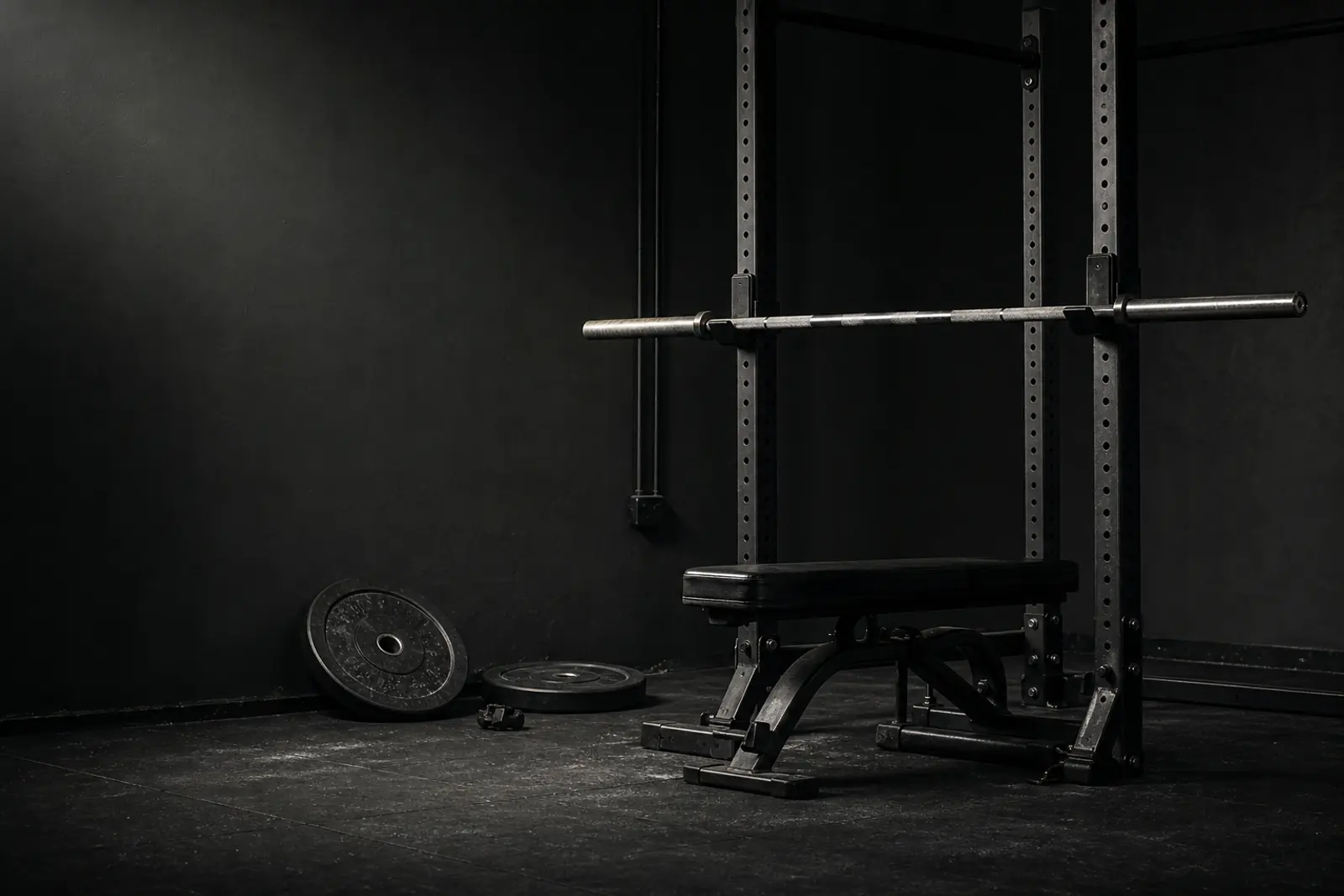 A quiet graphite gym with a squat rack, bench, barbell, and plates ready for a session.